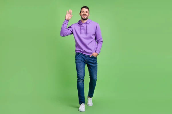 Full size φωτογραφία του αστείου Millennial brunet τύπος πάει κύμα φορούν hoodie τζιν sneakers απομονώνονται σε πράσινο φόντο — Φωτογραφία Αρχείου