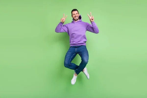 Full length photo of funky millennial brunet guy jump show v-sign wear hoodie jeans sneakers απομονωμένα σε πράσινο φόντο — Φωτογραφία Αρχείου