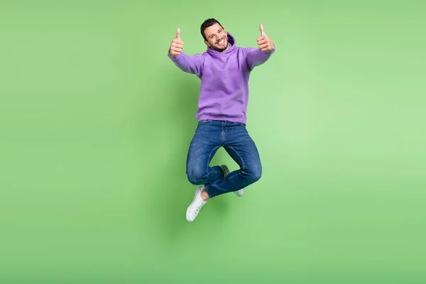 Полноразмерная фотография крутого молодого брюнета, прыгающего вверх большим пальцем в джинсах с капюшоном на зеленом фоне — стоковое фото
