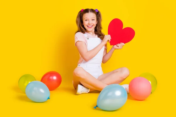 Plná délka tělo velikost fotografie školačka na party s balónky s úsměvem ukazující červené srdce izolované jasně žluté barvy pozadí — Stock fotografie