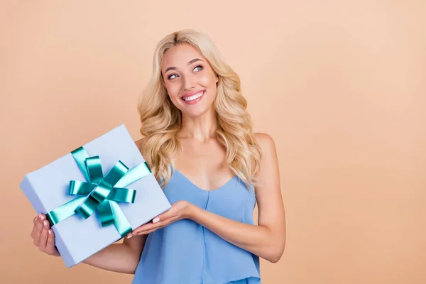 Photo of thoughtful sweet young lady wear blue singlet smiling holding gift box looking empty space isolated beige color background