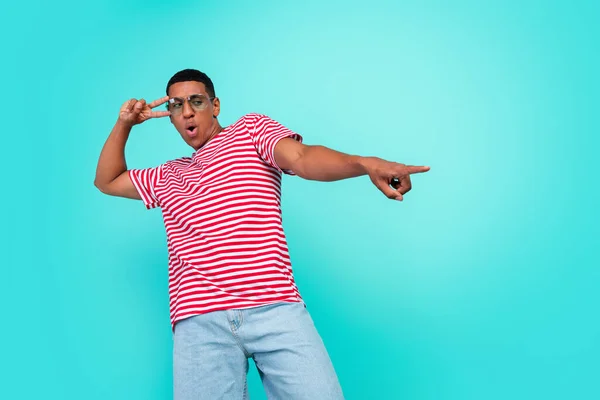Photo of excited cute young guy dressed red t-shirt dancing pointing empty space showing v-sign isolated turquoise color background — Stock Photo, Image