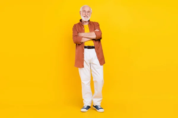 Fénykép vidám nyugdíjas öregember nagyapa keresztbe karok viselnek szemüveget barna ing nadrág cipő elszigetelt sárga színű háttér — Stock Fotó
