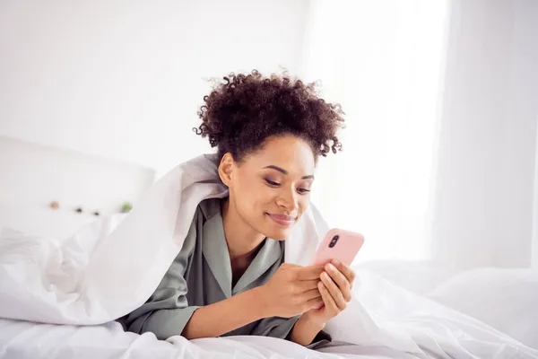 Portrait of attractive cheerful addicted focused wavy-haired girl lying in bed using gadget at light home house indoors