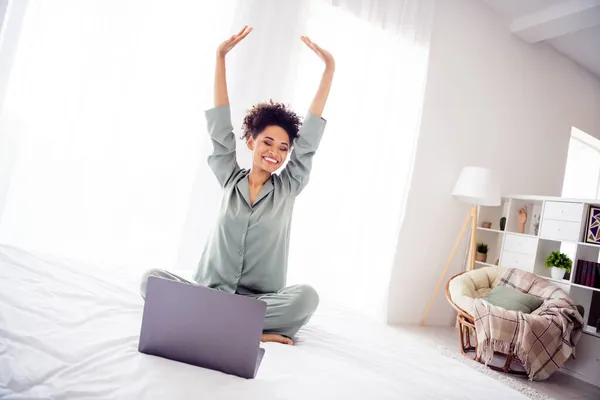 Retrato de chica alegre atractiva sentada en la cama usando el ordenador portátil que se extiende en el hogar blanco claro en el interior —  Fotos de Stock