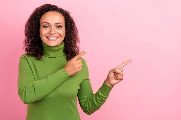 Foto de mulher afro muito brilhante usar gola alta verde apontando dedos espaço vazio sorrindo isolado cor de fundo rosa — Fotografia de Stock