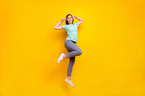 Foto in voller Größe von verrückten Brünette Frisur Millennial Dame springen V-Zeichen tragen T-Shirt Jeans Turnschuhe isoliert auf gelbem Hintergrund — Stockfoto