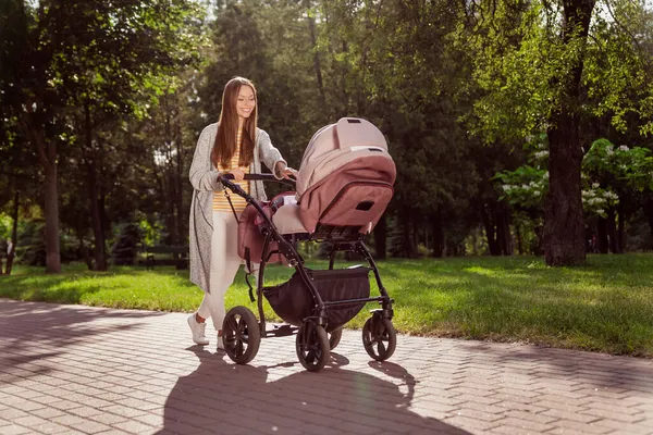 Longitud completa tamaño corporal foto joven madre sonriendo va en el parque de la ciudad con carro de bebé feliz —  Fotos de Stock