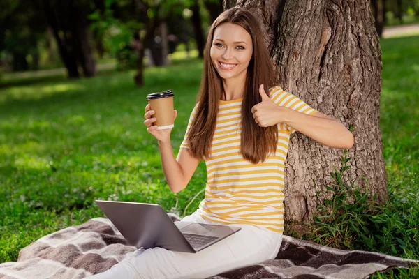 Retrato de menina alegre atraente sentado no gramado usando laptop beber café mostrando thumbup ao ar livre — Fotografia de Stock