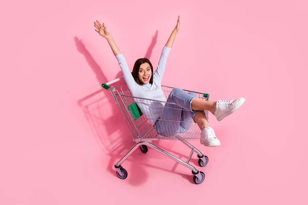Fénykép bájos vicces fiatal nő viselni fehér pulóver lovaglás bevásárlókocsi emelkedő karok mosolygós elszigetelt rózsaszín szín háttér — Stock Fotó