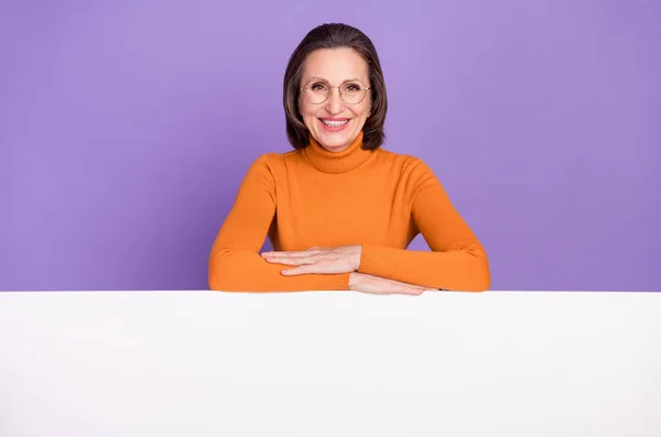 自信を持ってかなり女性の年金受給者の写真は、白い看板の空のスペースを保持オレンジ色のタートルネックメガネを身に着けている孤立紫色の背景 — ストック写真