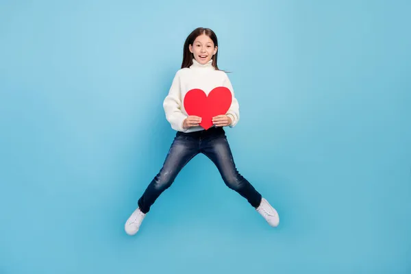 Foto de bonito adorável estudante menina desgaste branco suéter sorrindo salto alto segurando grande coração vermelho isolado azul cor fundo — Fotografia de Stock