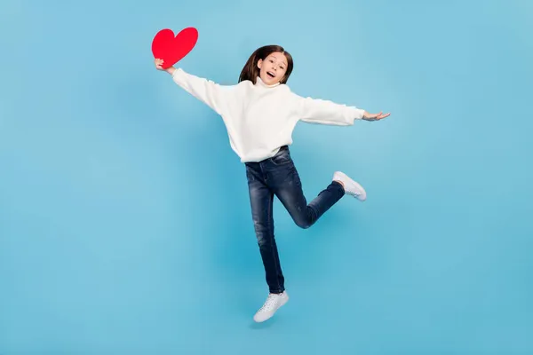 Foto de divertida chica preadolescente funky vestida de jersey blanco caminando saltando alto sosteniendo gran corazón rojo sonriendo aislado fondo de color azul — Foto de Stock