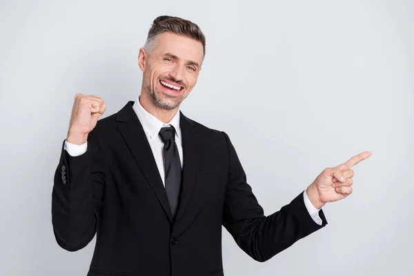 Фото счастливчика молодого менеджера носить черный пиджак поднимая кулак указывая пальцем пустое пространство изолированный серый цвет фона — стоковое фото