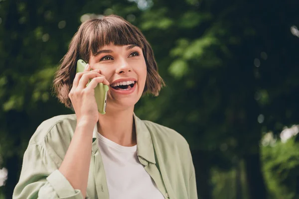 밝게 빛나는 웃는 젊은 아가씨가 녹색 셔츠를 입고 걸어 다니는 사진, 바깥 도시 공원에서 웃으며 현대식 가젯을 전달하는 모습 — 스톡 사진