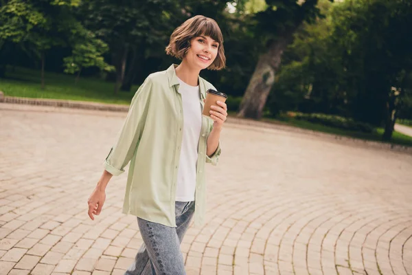 Фото милой очаровательной молодой леди в зеленой рубашке, идущей пить напиток улыбаясь на открытом воздухе городского парка — стоковое фото