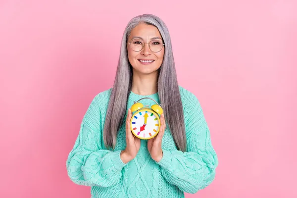Foto de bonito peinado blanco señora de edad mantenga reloj desgaste gafas de color verde azulado suéter aislado sobre fondo de color rosa — Foto de Stock