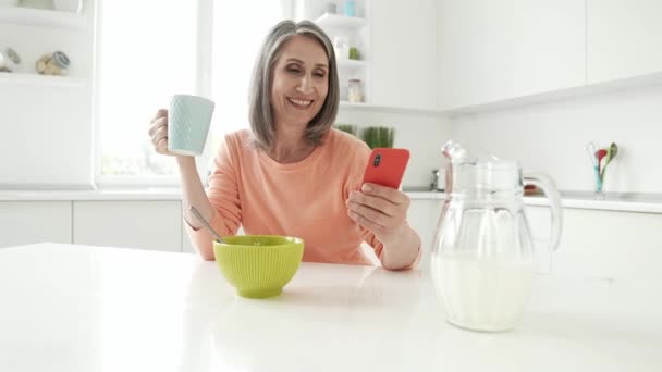 Positivo abuela hold teléfono escribir mensaje bebida sabroso cacao disfrutar del desayuno — Vídeo de stock