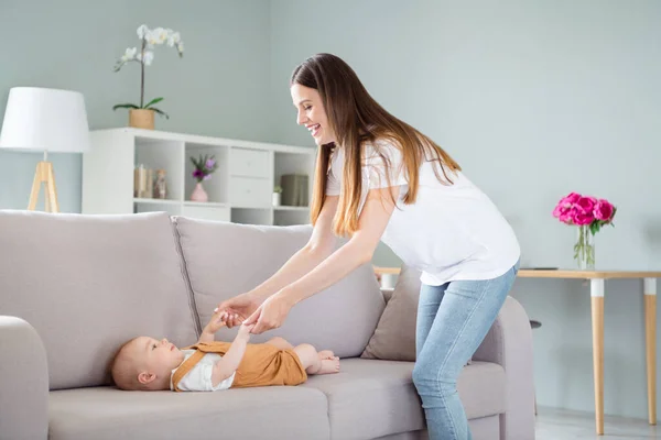 Foto di mamma carina infantile vestita di bianco t- tenendo le braccia bambino sdraiato divano sorridente all'interno della stanza appartamento — Foto Stock