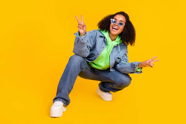 Foto de mujer afro dulce funky desgaste denim chaqueta brazo gafas oscuras sonriente sentado mostrando v-signos aislado color amarillo fondo —  Fotos de Stock