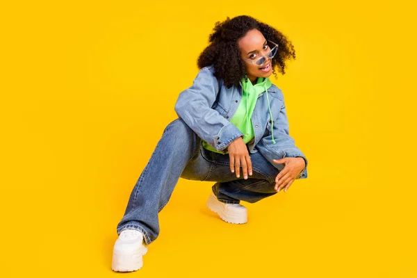 写真の魅力的な面白いアフリカの女性の服を着たジーンズグリーンパーカーダークアイウェア座って笑顔孤立した黄色の色の背景 — ストック写真