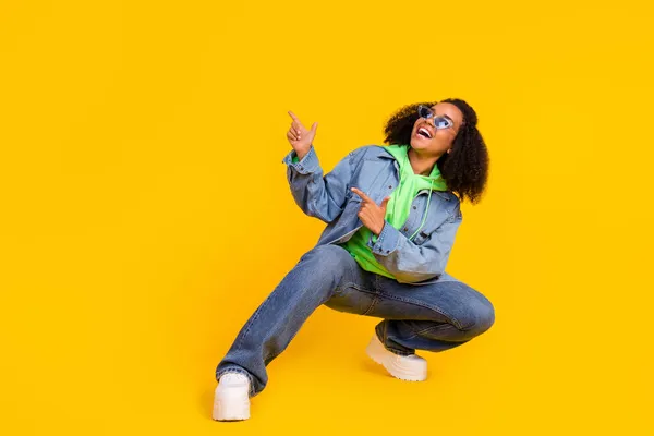 Foto de mujer africana bastante encantadora vestida jeans verde sudadera con capucha gafas oscuras señalando los dedos hacia arriba espacio vacío aislado color amarillo fondo — Foto de Stock