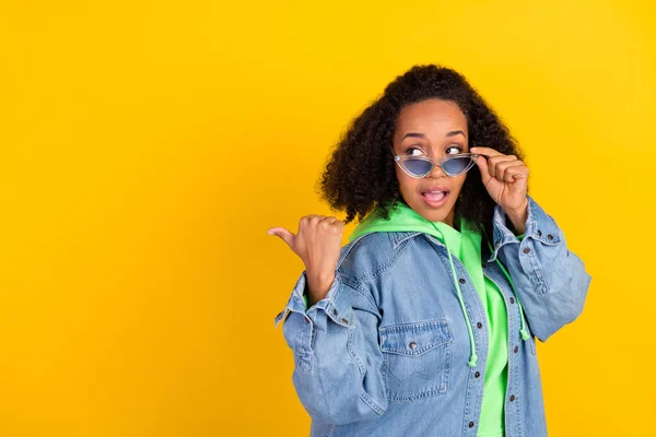 Foto de mujer afro hermosa atractiva usar denim chaqueta brazo gafas oscuras apuntando espacio vacío aislado color amarillo fondo —  Fotos de Stock