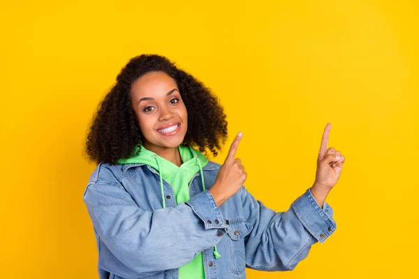 Φωτογραφία από γλυκό λαμπερό afro γυναίκα φορούν τζιν σακάκι δείχνοντας τα δάχτυλα μέχρι κενό χώρο χαμογελώντας απομονωμένο κίτρινο χρώμα φόντο — Φωτογραφία Αρχείου