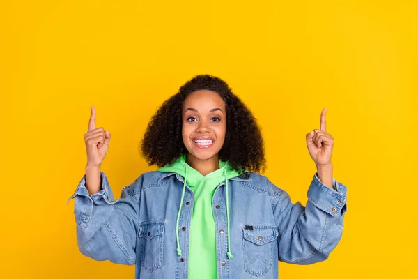 Foto van glanzend charmant afro vrouw dragen denim jas wijzen vingers omhoog lege ruimte glimlachen geïsoleerde gele kleur achtergrond — Stockfoto