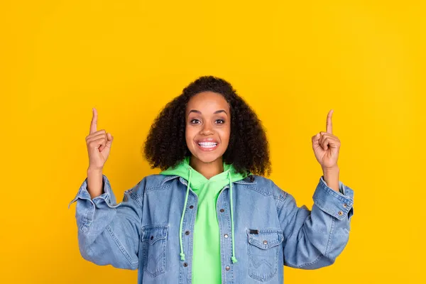 Foto av glänsande charmiga afro kvinna bära denim jacka peka fingrar upp tomt utrymme ler isolerad gul färg bakgrund — Stockfoto