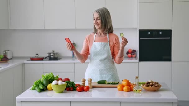 Blog yazarı etkisinde olgun bayan telefon parşömeni besleme ağı tutun ve salatalık yiyin — Stok video
