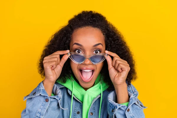 Bild av vackra imponerade afrikanska damen klädda jeans grön hoodie armar mörka glasögon ler öppen mun isolerad gul färg bakgrund — Stockfoto