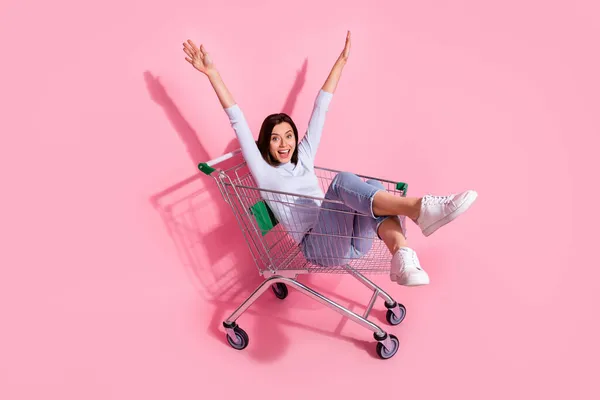 Foto van charmante grappige jonge vrouw dragen witte trui rijden winkelwagen rijzende armen glimlachen geïsoleerde roze kleur achtergrond — Stockfoto