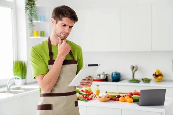 Портрет привабливого хлопця, який готує вечерю за допомогою пристрою, який дивиться веб-відео-підручник на домашній кухні в приміщенні — стокове фото
