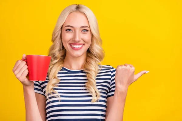 Portrett av en lystig jente som drikker cappuccino og viser reklameinnslag som ser isolert ut over livaktig gul bakgrunn – stockfoto