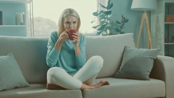 Pen pensjonert dame sitter med låra sofa og nyter morgencappuccino – stockvideo