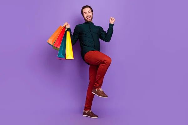 Фото смешного удачливого молодого парня одетого смарт-повседневной одежды, идущего поднимаясь кулаком по магазинам улыбаясь изолированный фиолетовый цвет фона — стоковое фото