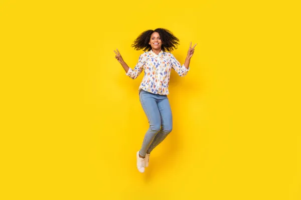 Foto em tamanho completo de muito jovem ondulado penteado senhora salto mostrar v-sign desgaste blusa jeans tênis isolado no fundo amarelo — Fotografia de Stock