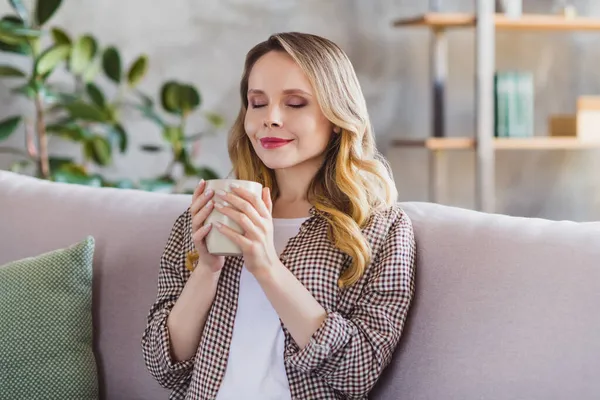 Фото милой блестящей молодой женщины, одетой в клетчатую рубашку, сидящей на диване и пьющей кофе. — стоковое фото