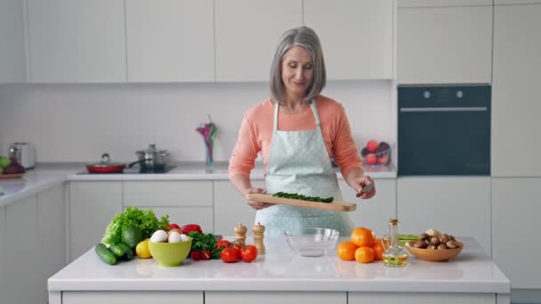 Wesoła babcia gospodyni domowa przygotować mieszankę sałatek dodać posiekany ogórek — Wideo stockowe