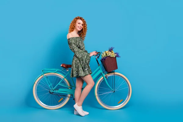 Volledige lengte profiel foto van schattige dame rijden fiets dragen groene jurk sneakers geïsoleerd op blauwe kleur achtergrond — Stockfoto