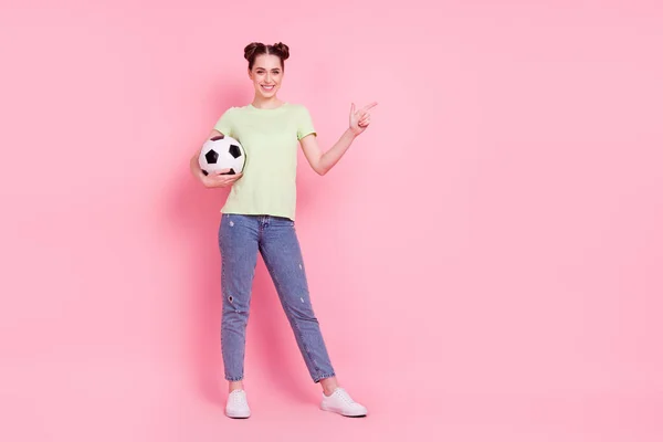 Fotografie de fată ține mingea de fotbal direct deget gol spațiu purta verde tricou blugi încălțăminte izolat pe fundal roz — Fotografie, imagine de stoc