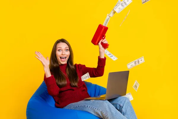 Φωτογραφία από κατάπληκτη μελαχρινή ευτυχισμένη νεαρή γυναίκα κατέχουν laptop sit καρέκλα χρήματα πτώση φορούν τζιν που απομονώνονται σε κίτρινο χρώμα φόντο — Φωτογραφία Αρχείου