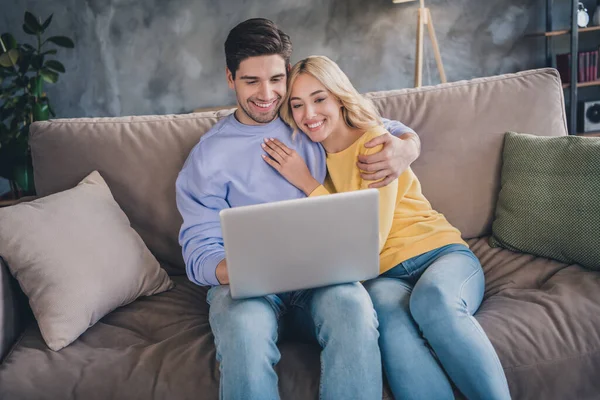 Foto de encantadores jovens amantes vestidos roupas casuais sofá sentado abraçando leitura gadget moderno sorrindo dentro de casa apartamento — Fotografia de Stock