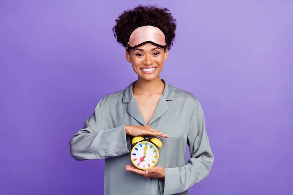 Φωτογραφία λαμπερό ελκυστική αφρικάνικη κυρία γκρι νυχτικά χαμογελώντας κρατώντας τα χέρια δείχνει ρολόι απομονώνονται μωβ φόντο χρώμα — Φωτογραφία Αρχείου