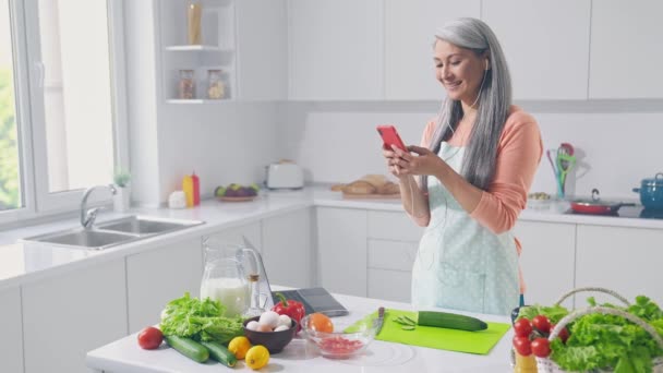 中年女性リッスンオーディオヘッドセットスマートフォン作るビーガン料理でキッチン — ストック動画