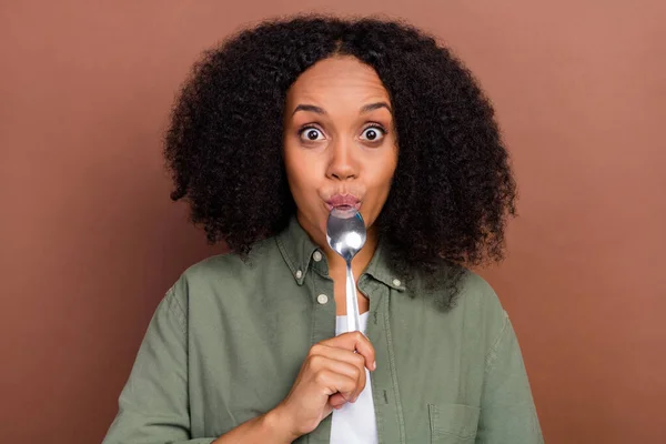 Foto de impressionado morena millennial afro penteado senhora lamber colher desgaste camisa cáqui isolado no fundo de cor marrom — Fotografia de Stock