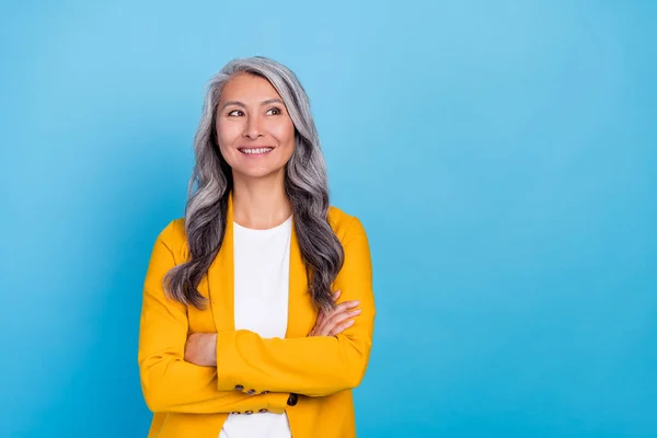 Foto de la señora mayor encantadora soñadora vestida con brazos de chaqueta amarilla plegada mirando espacio vacío sonriendo aislado color azul fondo — Foto de Stock