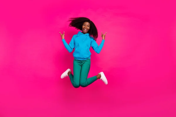 Фото фанки довольно темная кожа леди носить синий капюшон прыгая высоко показывая жесткий рок знак улыбаясь изолированный розовый цвет фона — стоковое фото