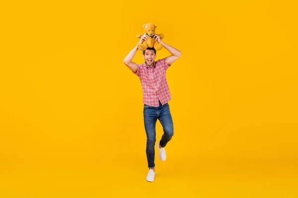 Pełna długość zdjęcie wesoły młody człowiek trzymać niedźwiedź zabawka nad głową chodzić izolowane na żółtym tle kolor — Zdjęcie stockowe